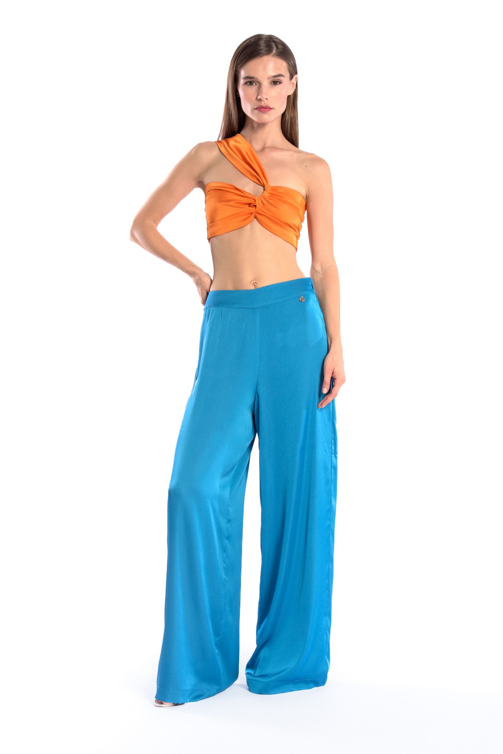 Model wearing a silk cross shoulder crop top in orange with back firefly logo golden zipper and silk pants in blue color with a firefly golden charm